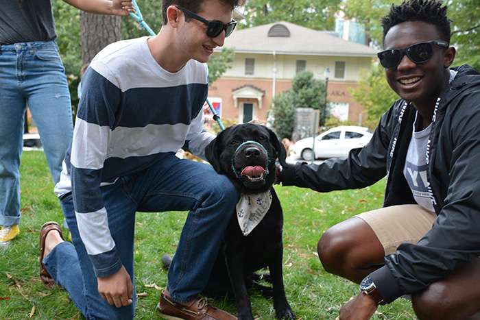 Two students in sunglasses kneel next to a black dog wearing a bandana