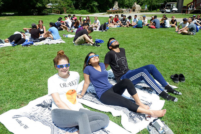 Several BBA students sit on a lawn in Clayton, Georgia, wearing solar eclipse-viewing glasses.