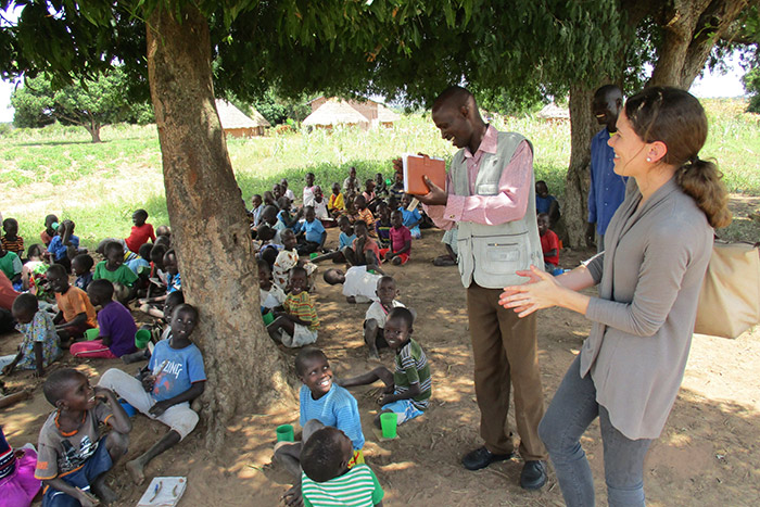 A large group of young Ugandans sit under a tree while two adults speak to them.