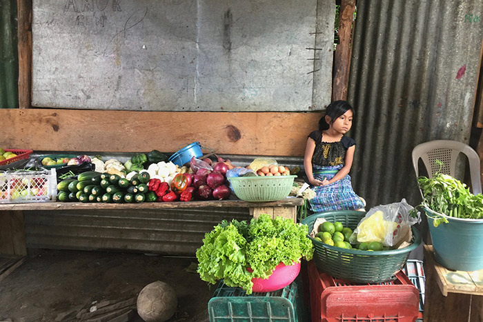 A young Guatemalan girl looks off in the distance, sitting on a bench filled of produce.