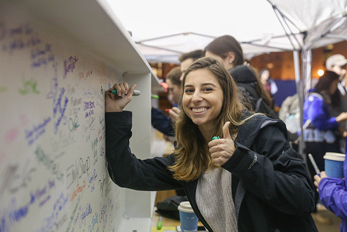 A student adds her name in green marker while giving a "thumbs-up" to the camera.