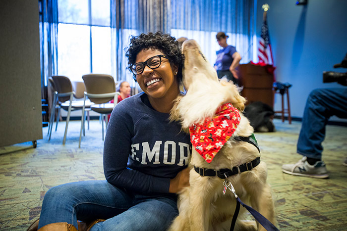 A student in an Emory sweatshirt sits on the ground with a huge smile on her face as she hugs an golden retriever therapy dog.