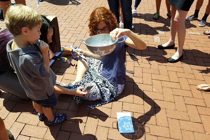Woman holds colander up for child to see eclipse crescents. 