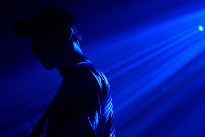 A member of the group stands in the dark blue hue of stage lighting.