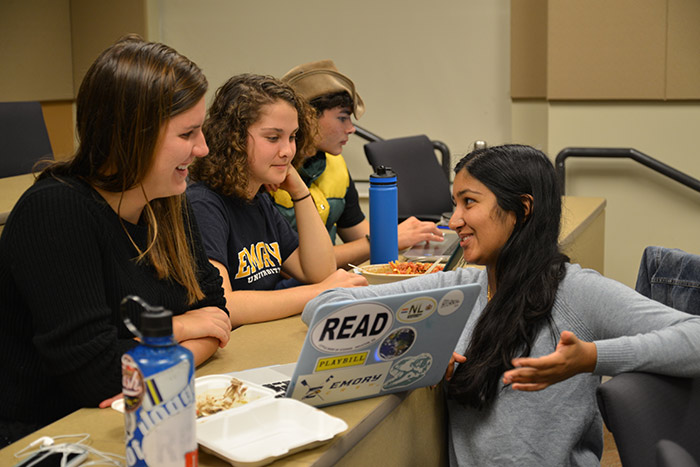 Three young women sit in a row in front of laptops while Maya Nair kneels in front of their table, pointing to something on a computer screen.