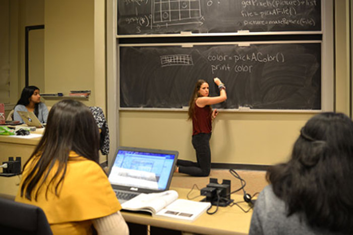 Tammany Grant kneels in front of a chalkboard at the front of a classroom, while young women take notes as she speaks and adds notes to the chalkboard.