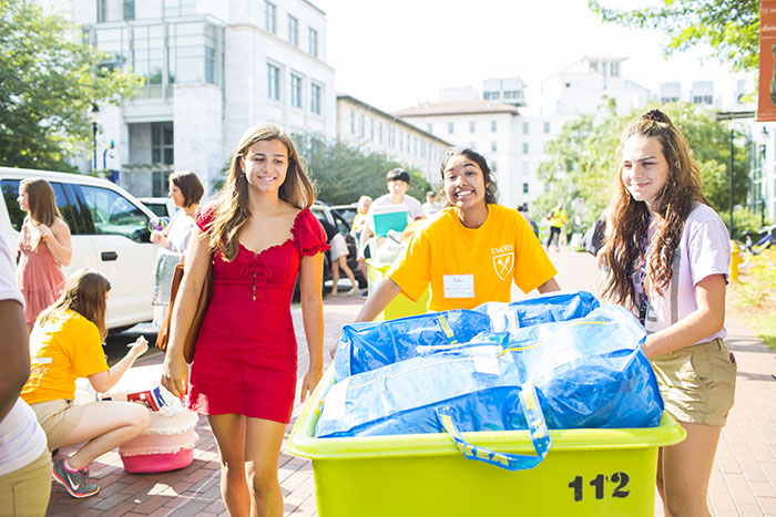 A student volunteer in a yellow t-shirt helps two students move in by rolling items in a large cart