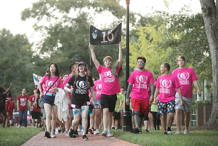 Students wearing pink t-shirts follow behind a teammate carrying a sign