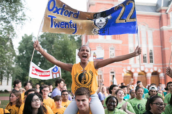 A student wearing an Emory gold t-shirt carries a banner while riding on the shoulders of another classmate