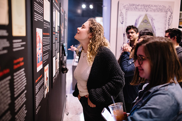 Two young women and several other students in the background read exhibit information hung on the wall