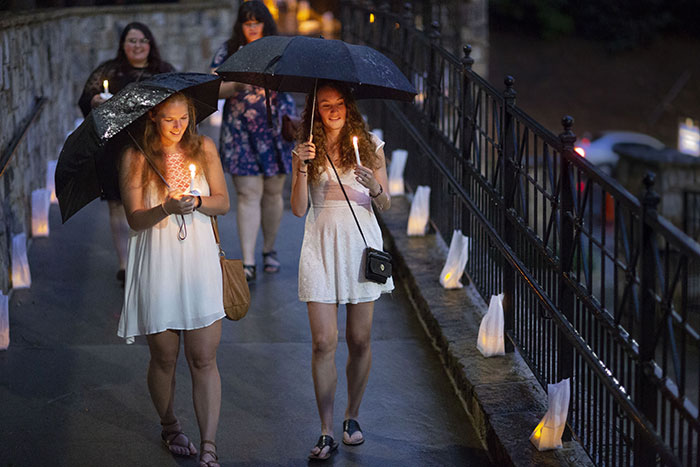 Two young women carrying umbrellas and wearing white dresses hold candles as they cross the bridge