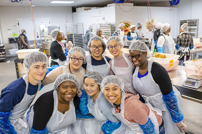 A group of students wear hair nets and aprons as they prepare meals at Project Open Hand