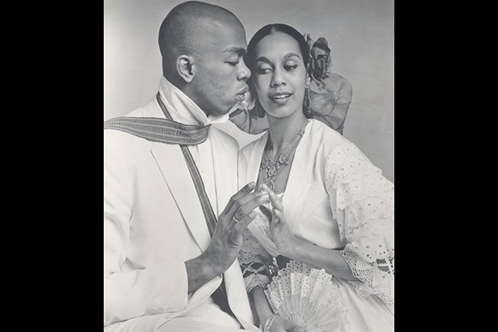 Carmen de Lavallade and Geoffrey Holder pose for a photo, sitting side-by-side, their fingertips touching each other as they look at each others lips.