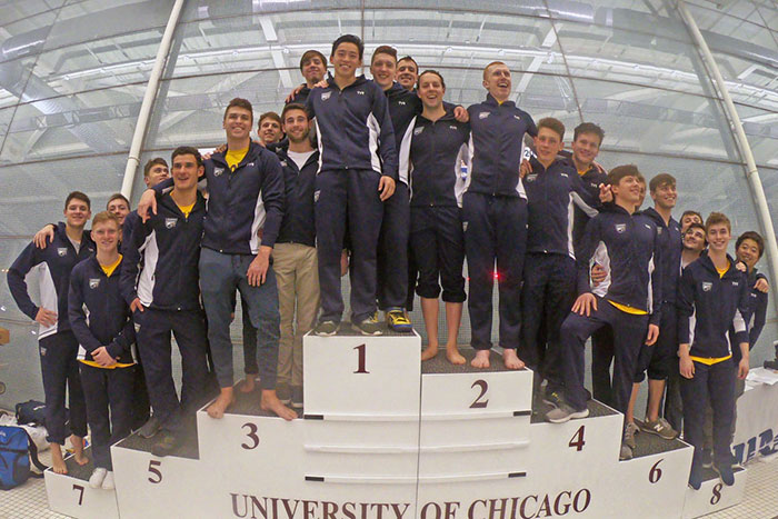 The Emory men's Swimming & Diving team stands on a riser together, arms around each other, posing for a champions photo in matching Emory Athletics jackets