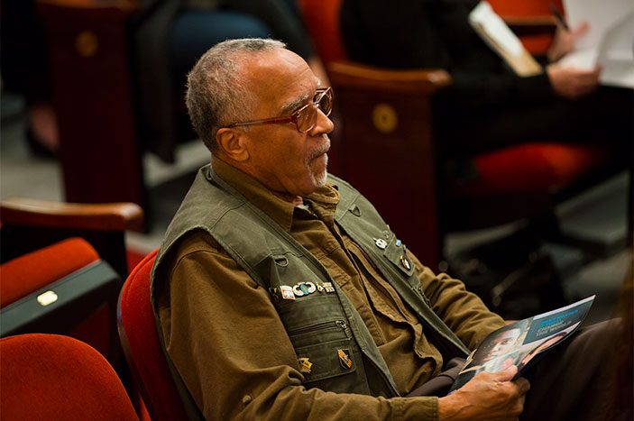 A man wearing a vest with military pins sits in the audience