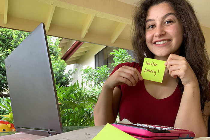 A student sits in front of her laptop and holds up a post-it that says 'Hello Emory'