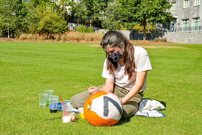 A young woman wearing a mask paints a pumpkin on the quadrangle