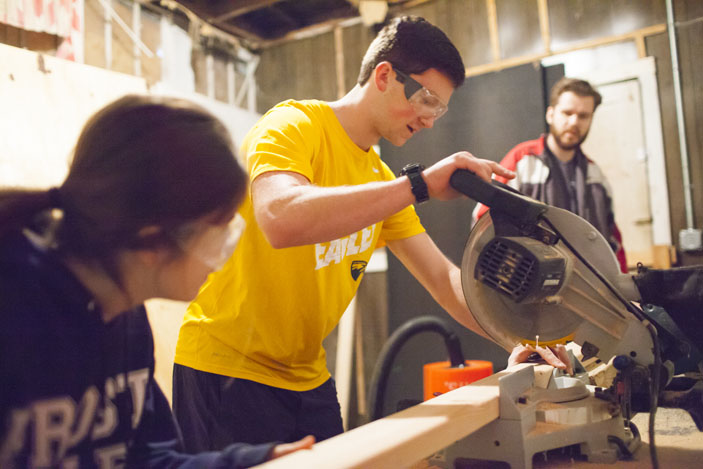 A volunteer in bright yellow Emory Cares shirt operates a table saw while others look on.