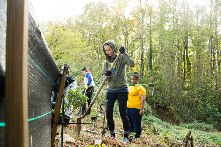 A female student volunteer in a hooded sweatshirt digs with a shovel along a silt barrier fence. Another volunteer looks on.