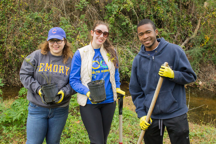 Three volunteers pose with shovels and potted plants, which they are about to plant.
