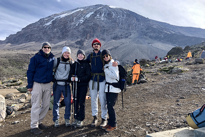 The group of hikers stand in front of their camp site with Mount Kilimanjaro in the distance