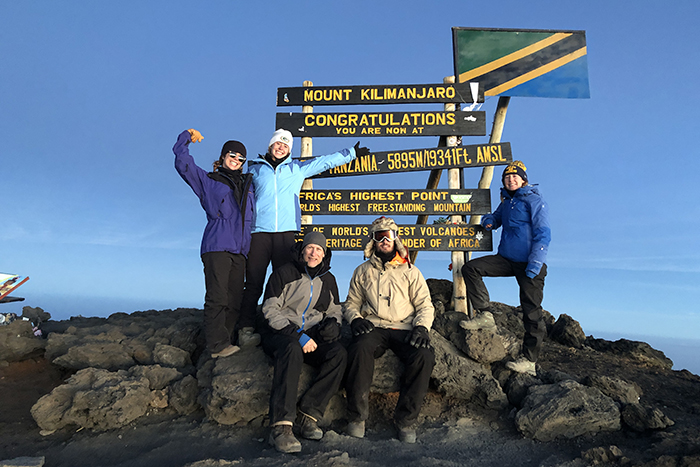 Five people stand at the Mount Kilimanjaro summit in front of a sign congratulating them