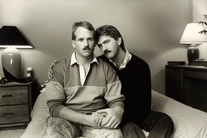 Two men sitting on a bed, one with an arm around the other