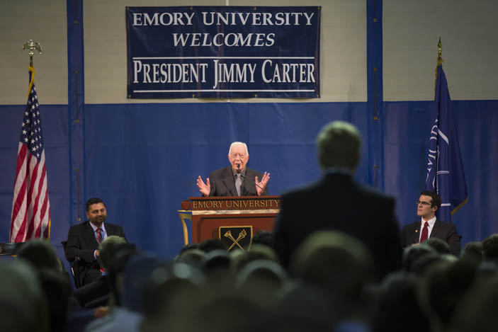 President Carter speaking at the podium, both hands raised in a gesture. In the background a banner says, "Emory University Welcomes President Jimmy Carter."