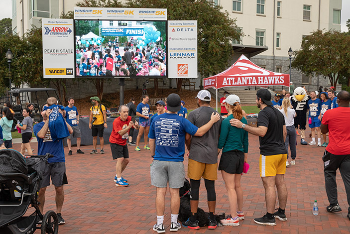 People at event standing near big screen showing runners and walking coming in